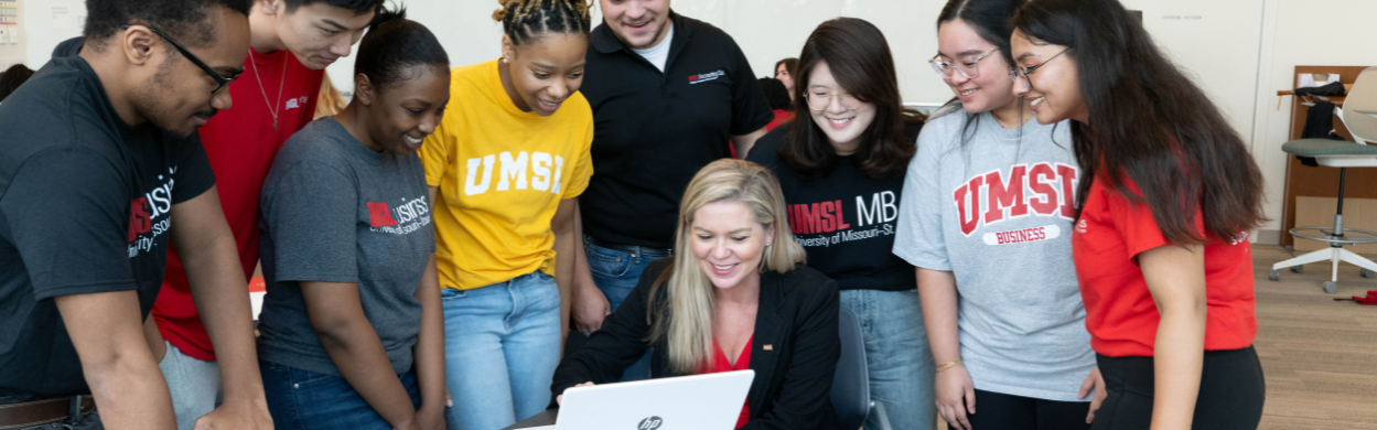 Dr. Cindy Goodwin-Sak sitting at a table with a laptop surrounded by students in UMSL gear