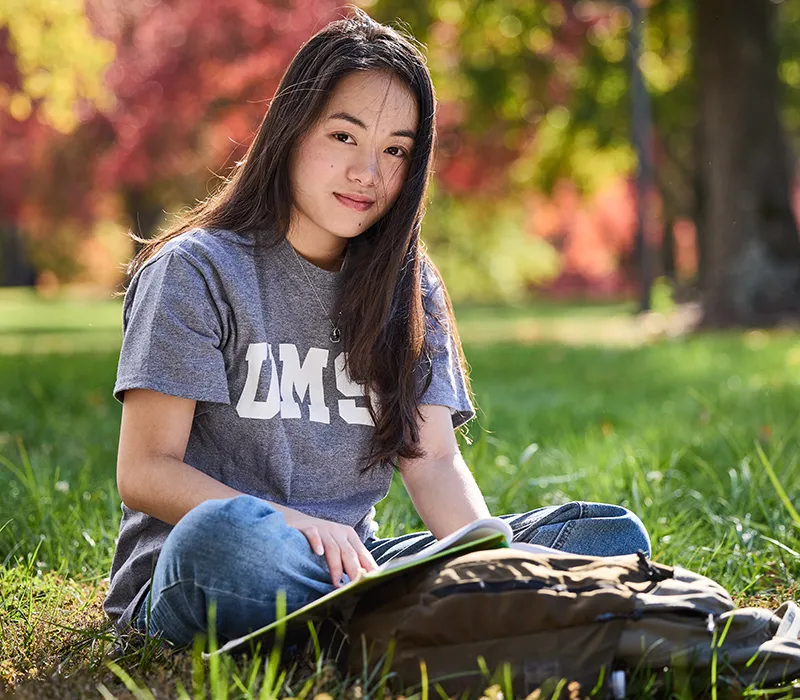 Student sitting on grass with open book and backpack