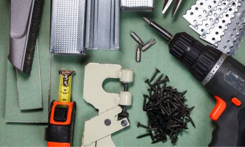 A variety of construction tools and supplies, including a power drill, a tape measure, drywall screws, and edge trim.