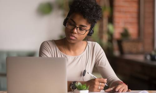 A young woman wearing headphones and seated at a laptop, holding a pen and poised to take notes.